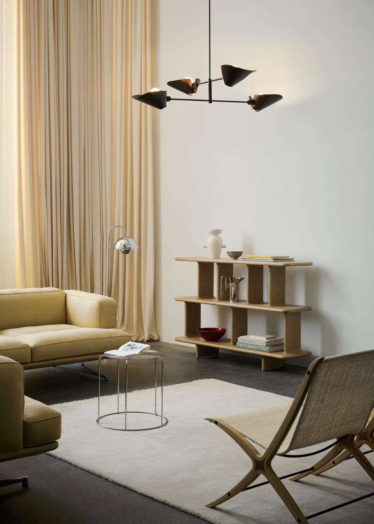 A bright living space features a light yellow sofa and armchair, a chrome Flowerpot VP12 floor lamp, and a natural wood tiered shelf with decor. A light rug covers a dark floor, while large windows are draped with light-colored curtains. A dark, multi-bulb chandelier hangs above.