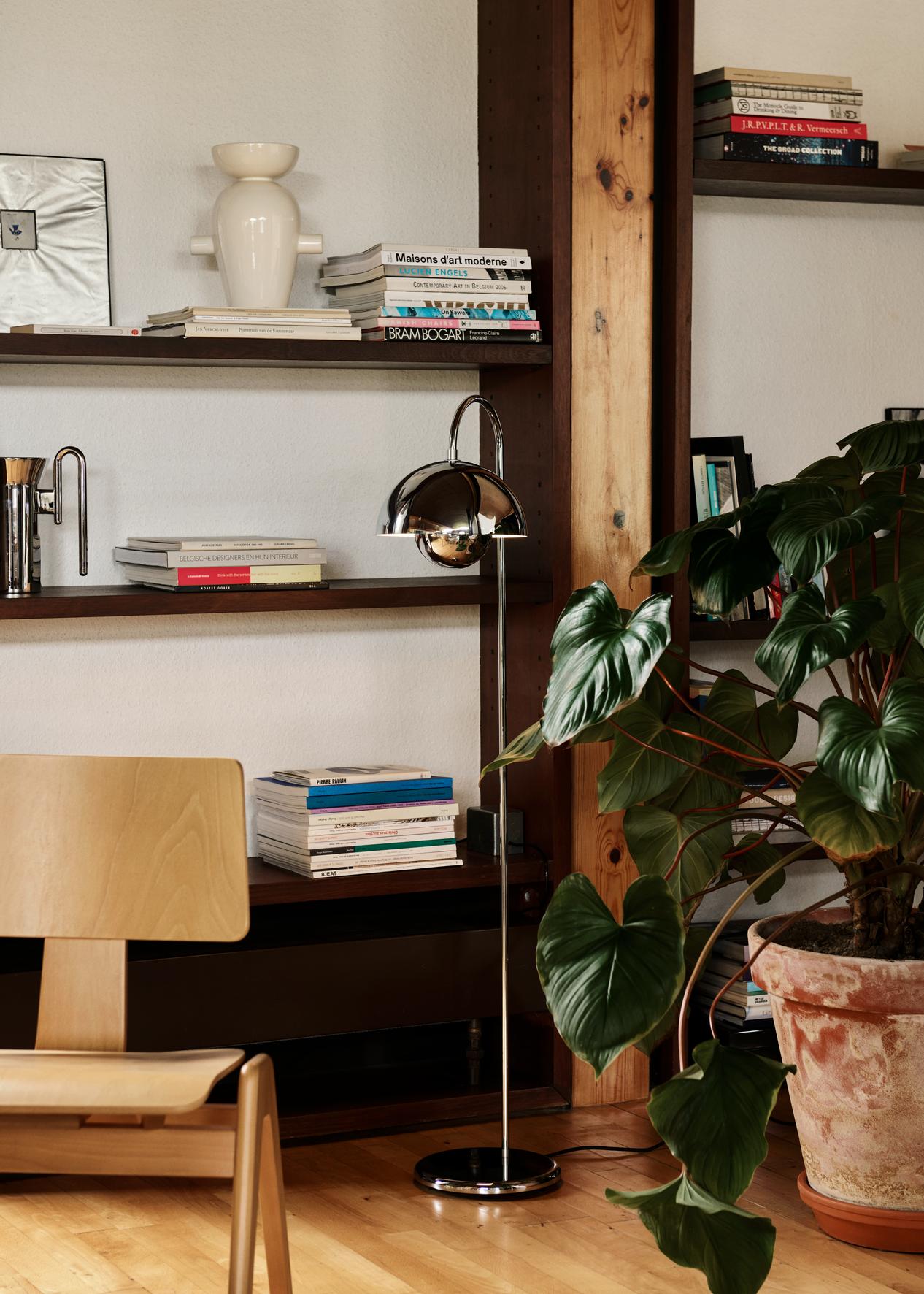 A polished chrome Flowerpot VP12 floor lamp stands in front of dark wooden shelves filled with books and decor. A light wood side chair is partially visible on the left, and a large potted plant sits to the right of the lamp.