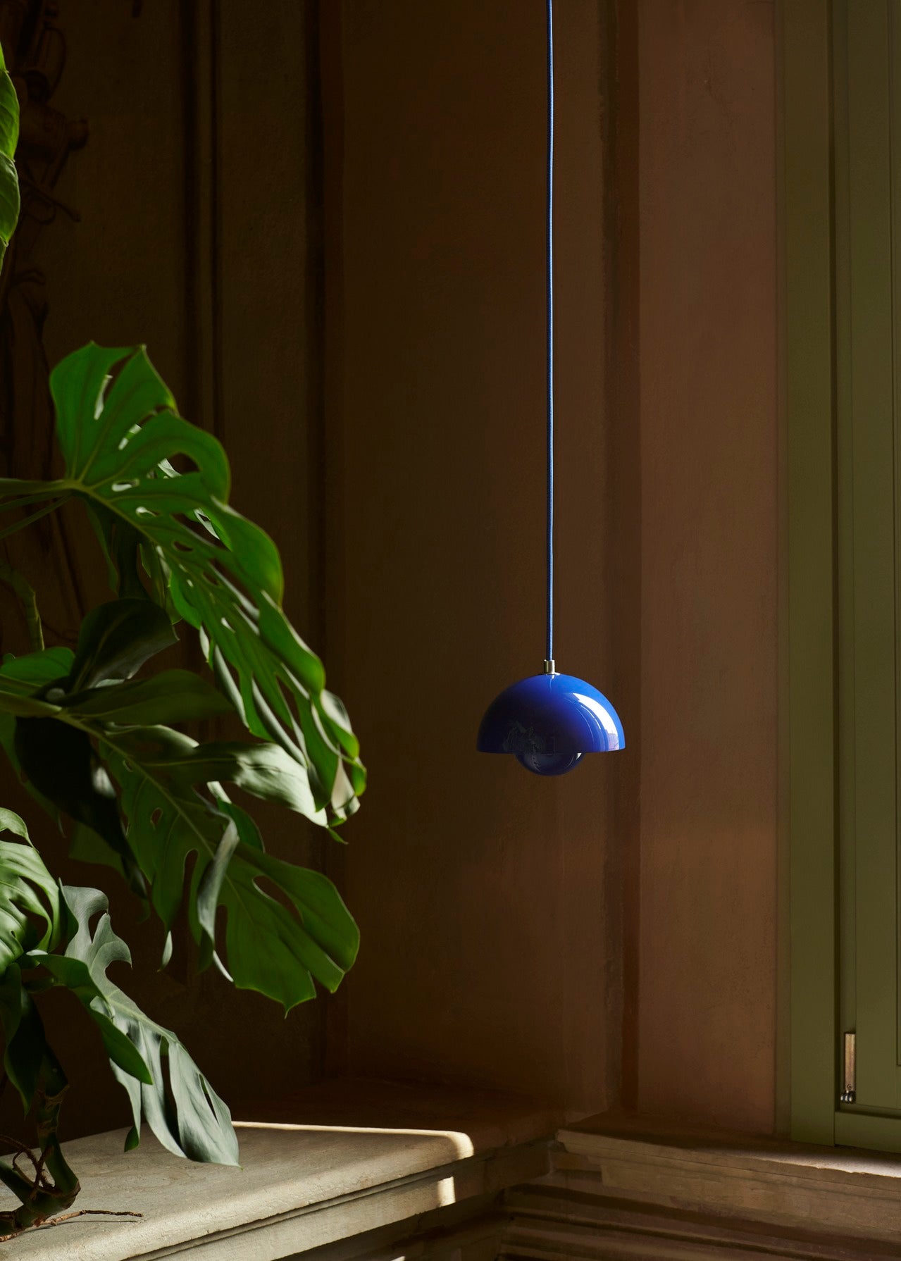 A vibrant, glossy, blue Flowerpot VP10 pendant lamp hangs from a thin cord against a warm background. To the left, large green leaves of a plant partially frame the image.