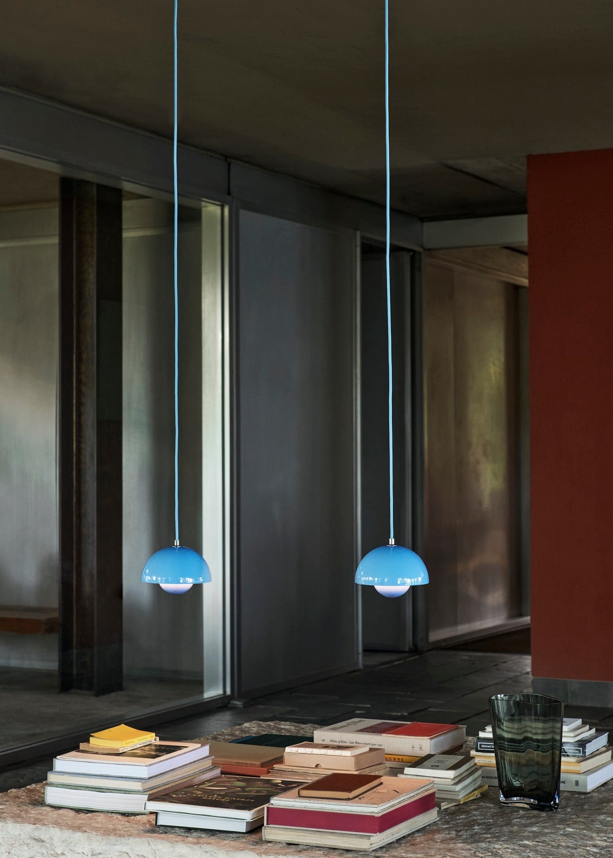 Two light blue Flowerpot VP10 pendant lamps hang from long, thin cords in a room with a dark ceiling and industrial elements. Below them, a collection of stacked books and a dark gray glass vase sit on a textured surface.