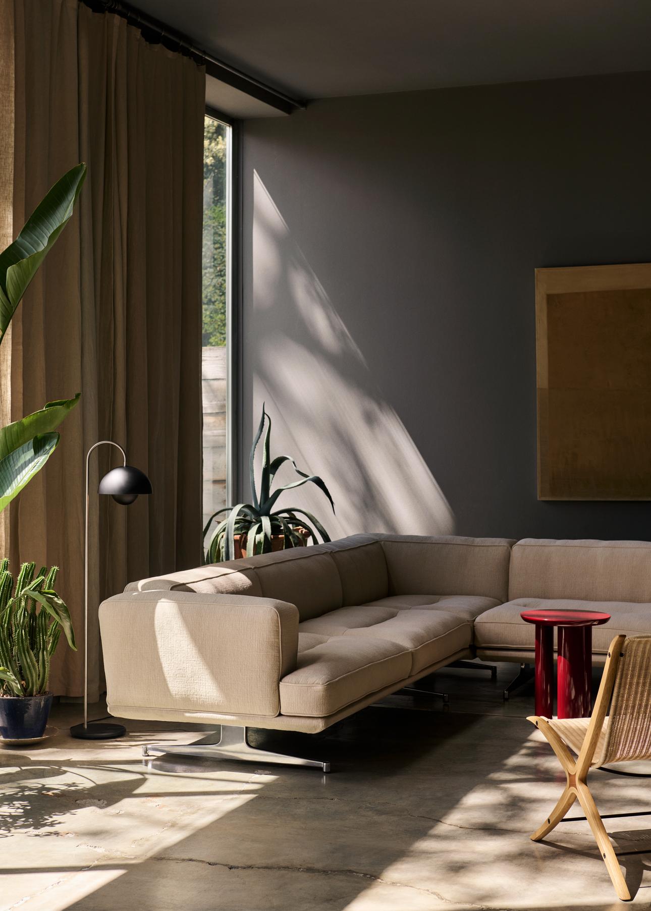 A modern, sunlit living room features a beige L-shaped sofa with a red side table. A black Flowerpot VP12 floor lamp and tall green plants are by a large window.