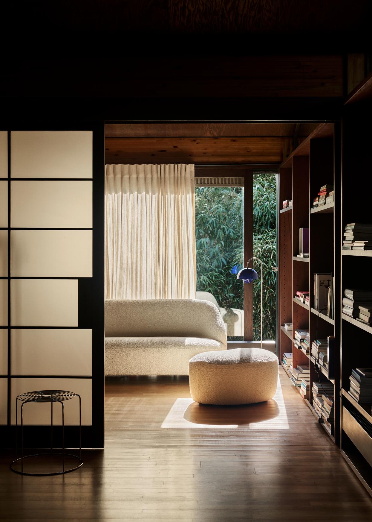 A warm, dark-paneled room with bookshelves opens to a brighter space featuring a creamy boucle sofa and ottoman, a cobalt blue Flowerpot VP12 lamp by a window with sheer curtains, and a view of greenery. A dark wire stool is in the foreground.