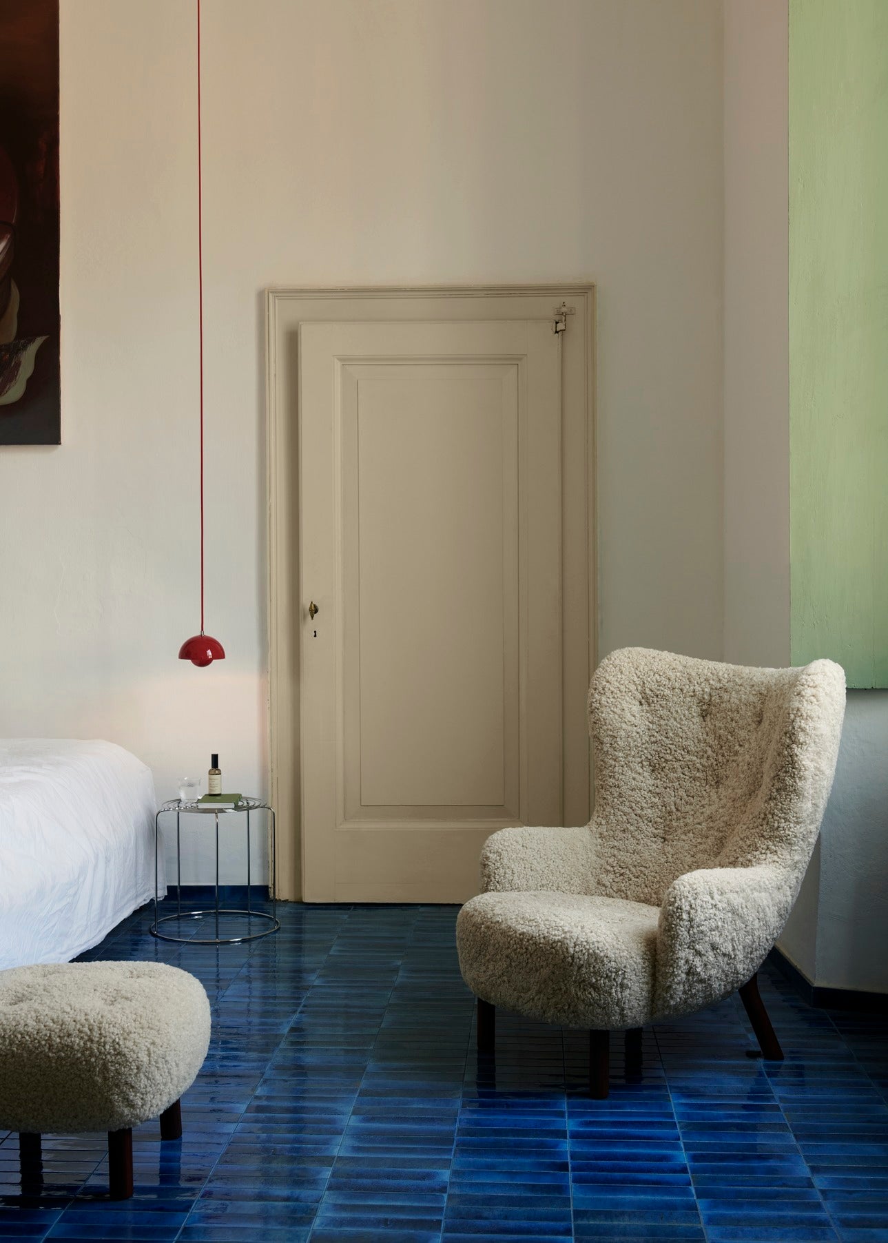 A modern room features blue tiled floors, a cream boucle armchair and pouf, and a wire stool beside a white bed, illuminated by a red Flowerpot VP10 pendant lamp.