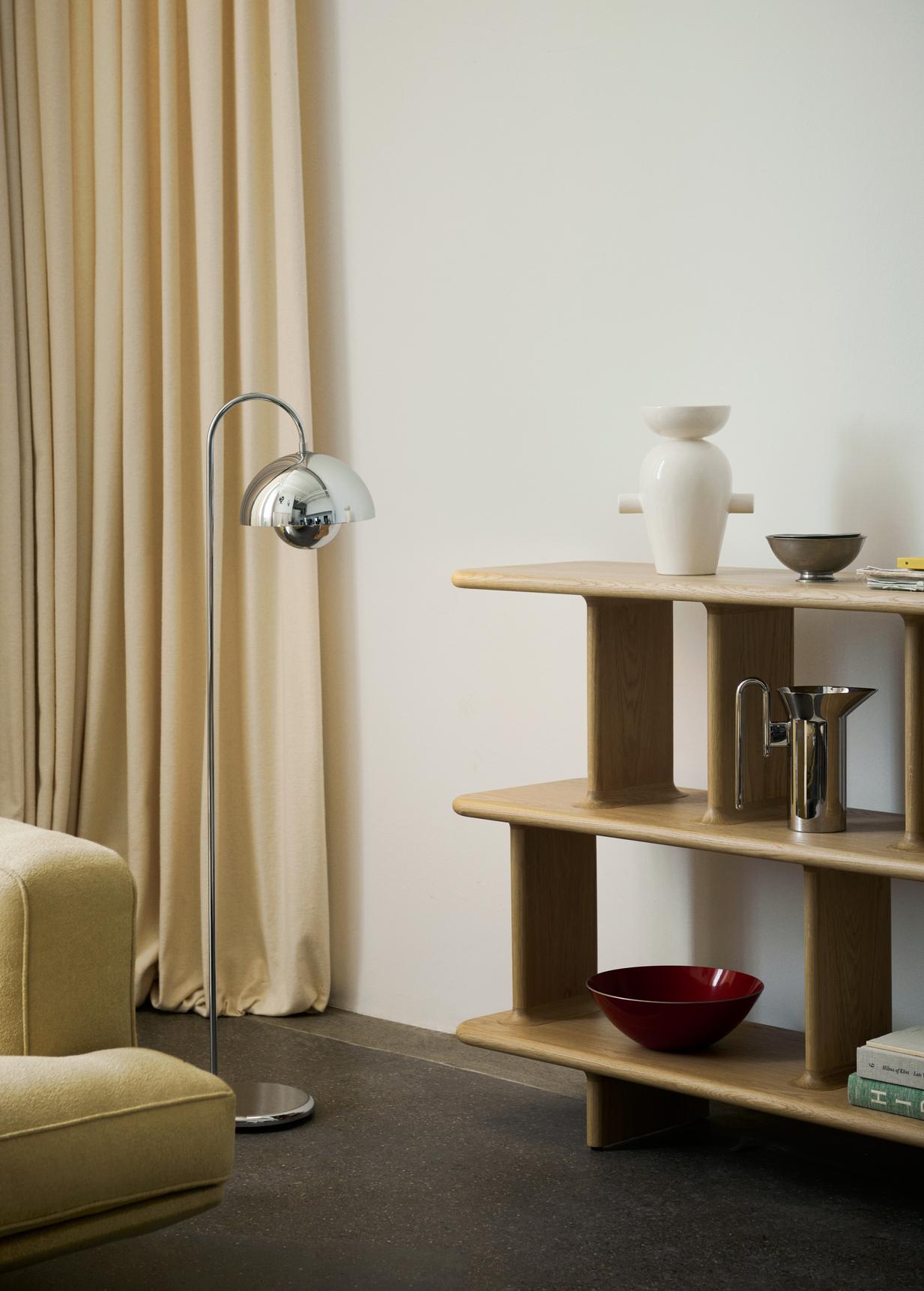 A bright interior shot features a chrome Flowerpot VP12 floor lamp, a light yellow sofa section, and a natural wood tiered shelf holding a white vase, bowls, and books. Light-colored curtains frame the left, and a dark floor completes the scene.
